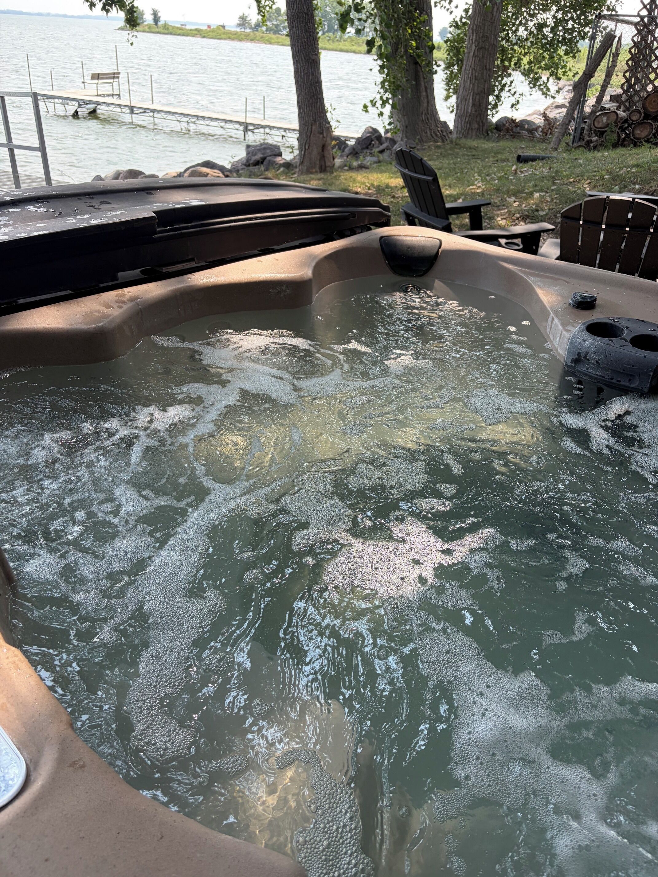 Ein Whirlpool mit Holzterrasse und Stühlen, umgeben von Bäumen und einem Gewässer.