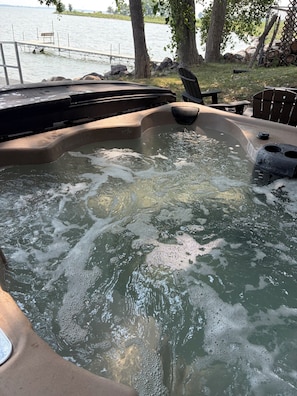 Ein Whirlpool mit Holzterrasse und Stühlen, umgeben von Bäumen und einem Gewässer.