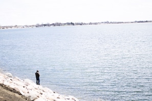 Une personne se tient près d’une étendue d’eau calme, avec une côte lointaine à l’horizon.