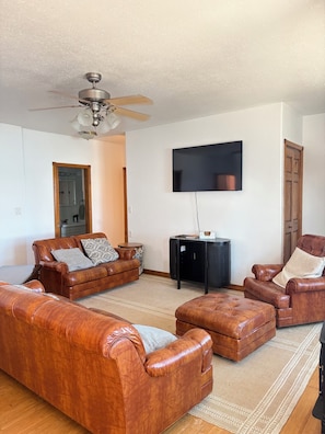 A living room with a ceiling fan, a flat-screen TV mounted on the wall, and leather sofas. - Cozy beach front lake cabin with hot tub! (Lake Norden)