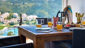 Ett träbord dukat med ett glas apelsinjuice, ett glas vitt vin och en kanna apelsinjuice. I bakgrunden syns en vacker utsikt över en stad och berg.