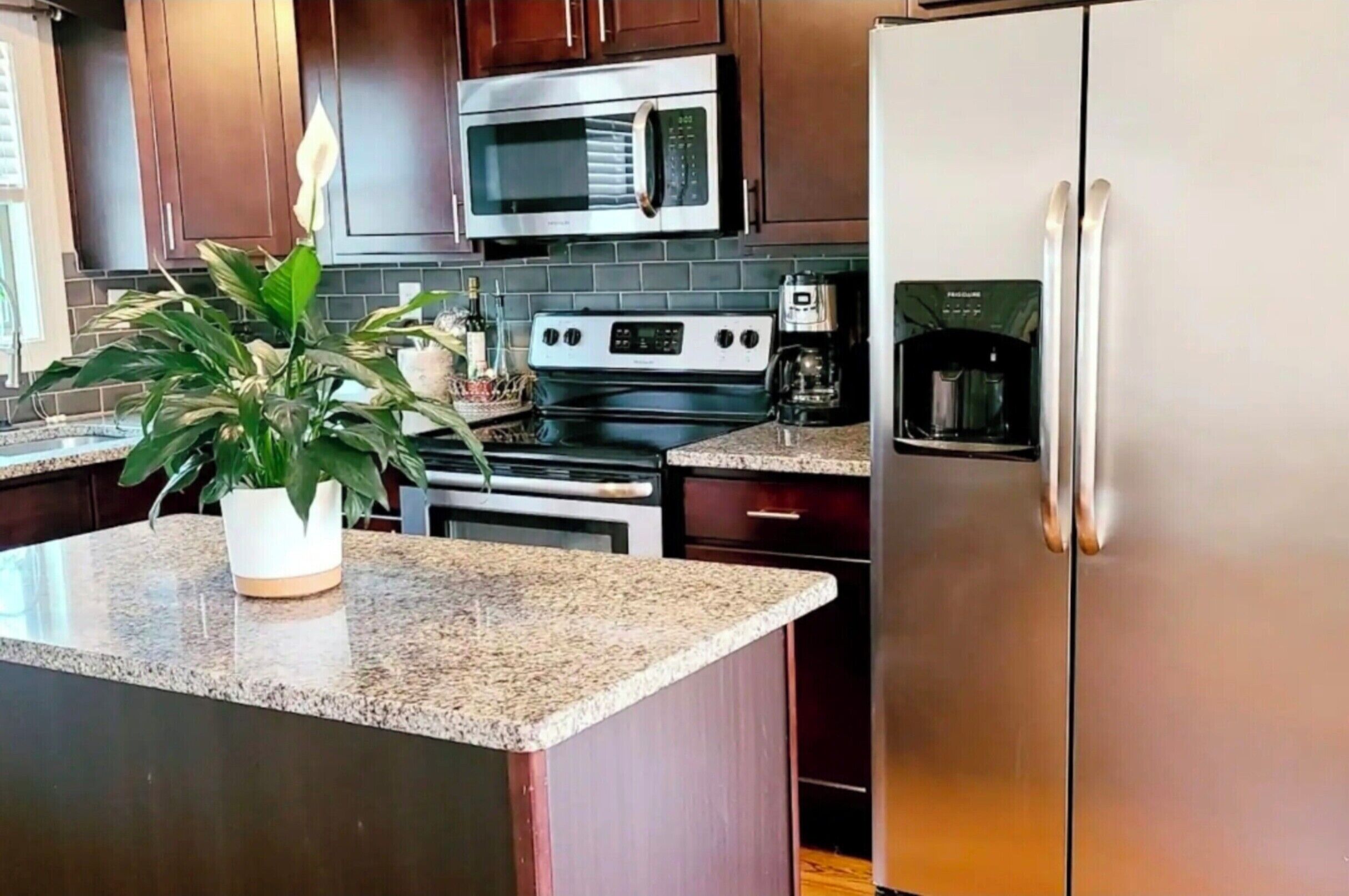 A kitchen with dark wooden cabinets, stainless steel appliances including a microwave, oven, and refrigerator, a granite countertop, and a potted plant.