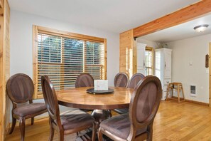 A dining area with a round wooden table and six chairs, large windows with blinds, and a white cabinet in the background.