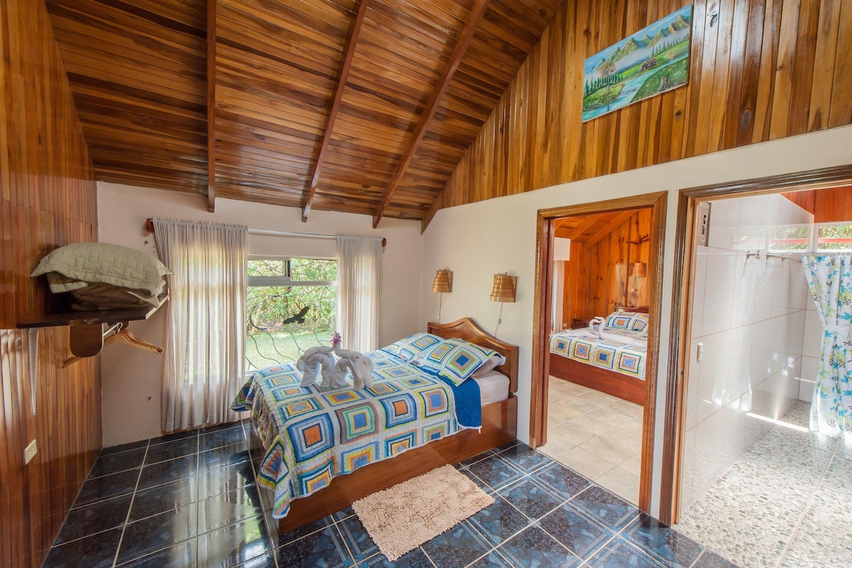 A bedroom with a wooden ceiling, a bed with a colorful bedspread, a window with curtains, and a view of greenery outside.