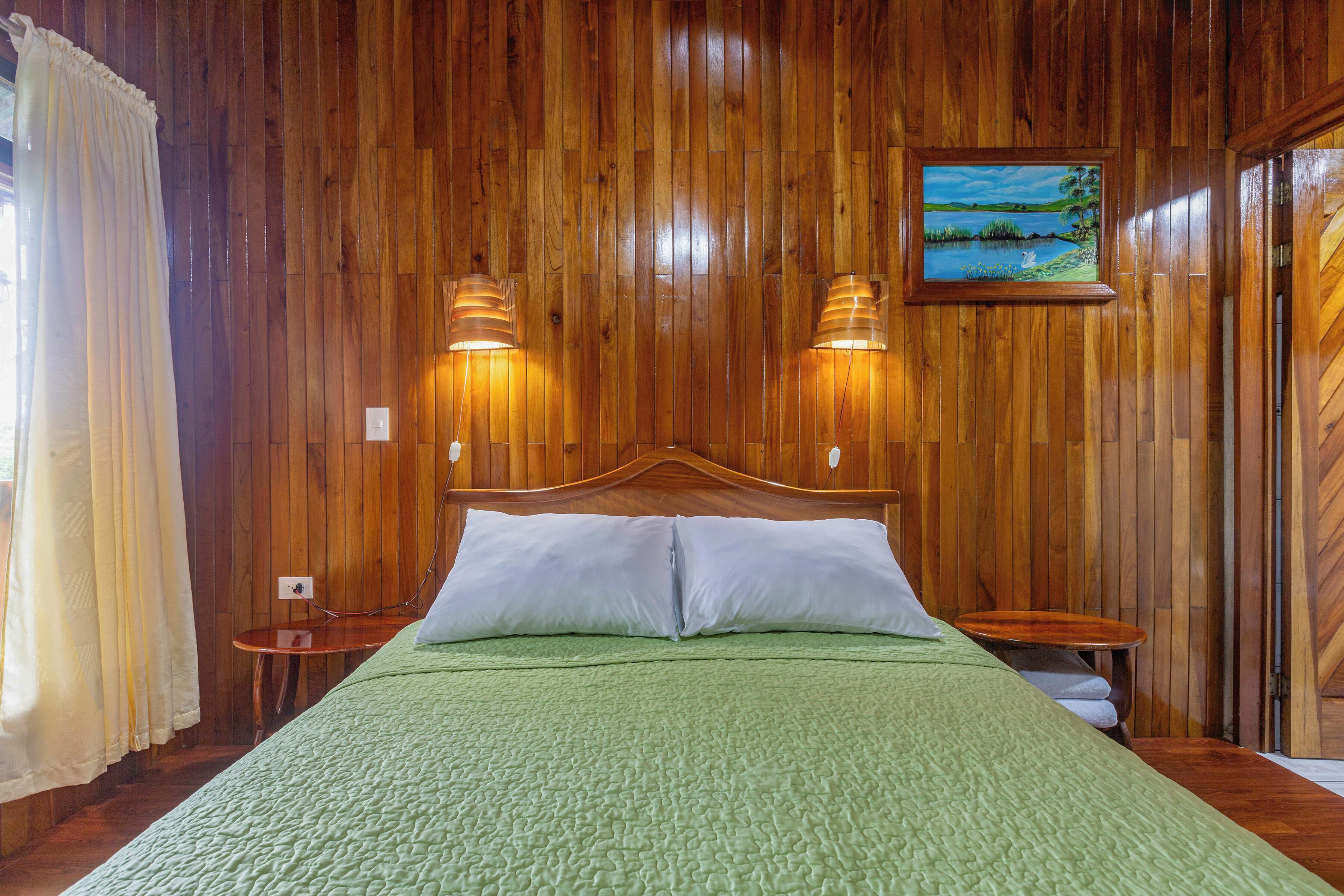 A wooden-paneled room with a bed, two bedside lamps, a painting, and a wooden door.