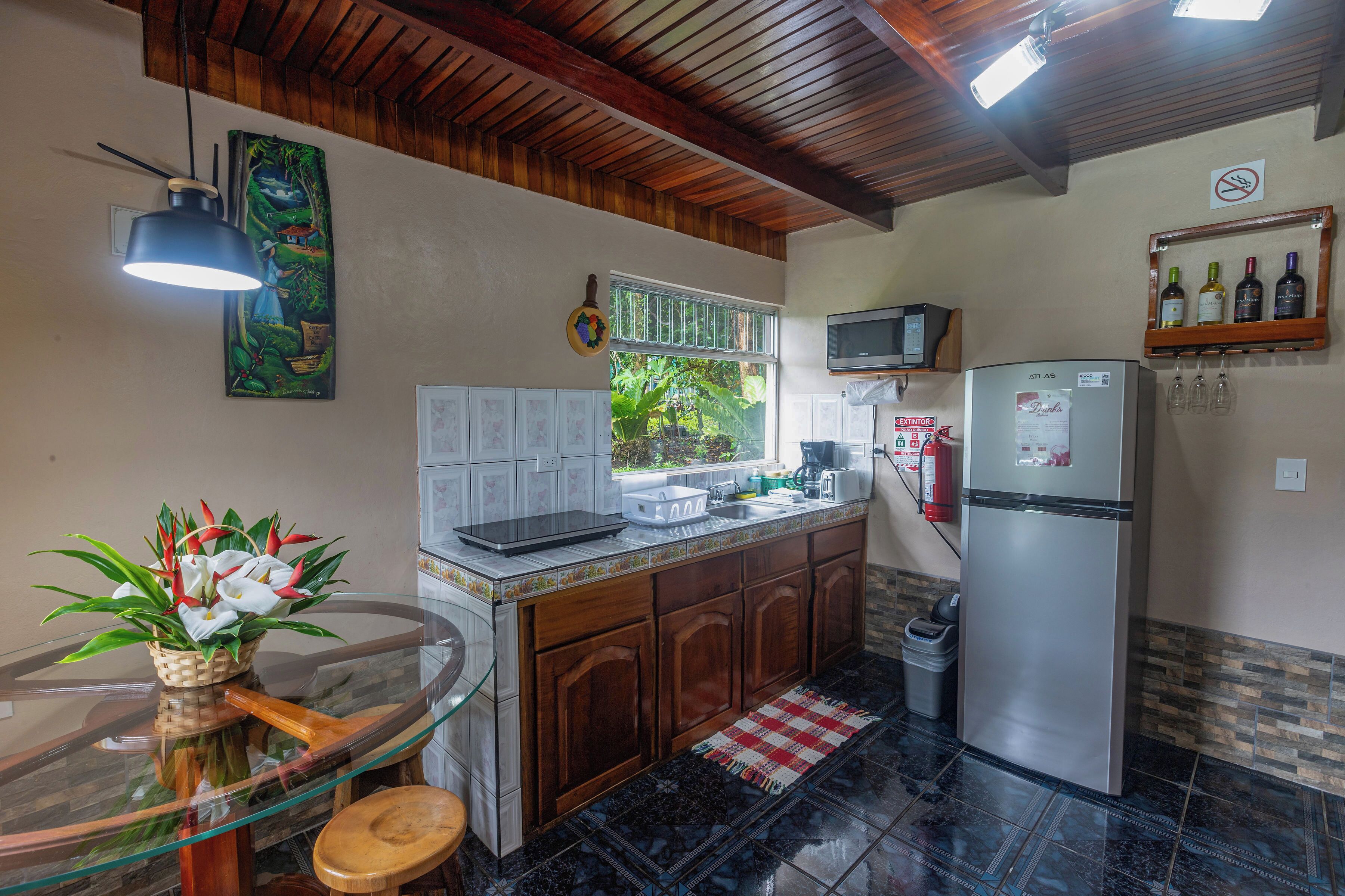 A kitchen with a wooden ceiling, a glass table, a refrigerator, and a window with a view of greenery.