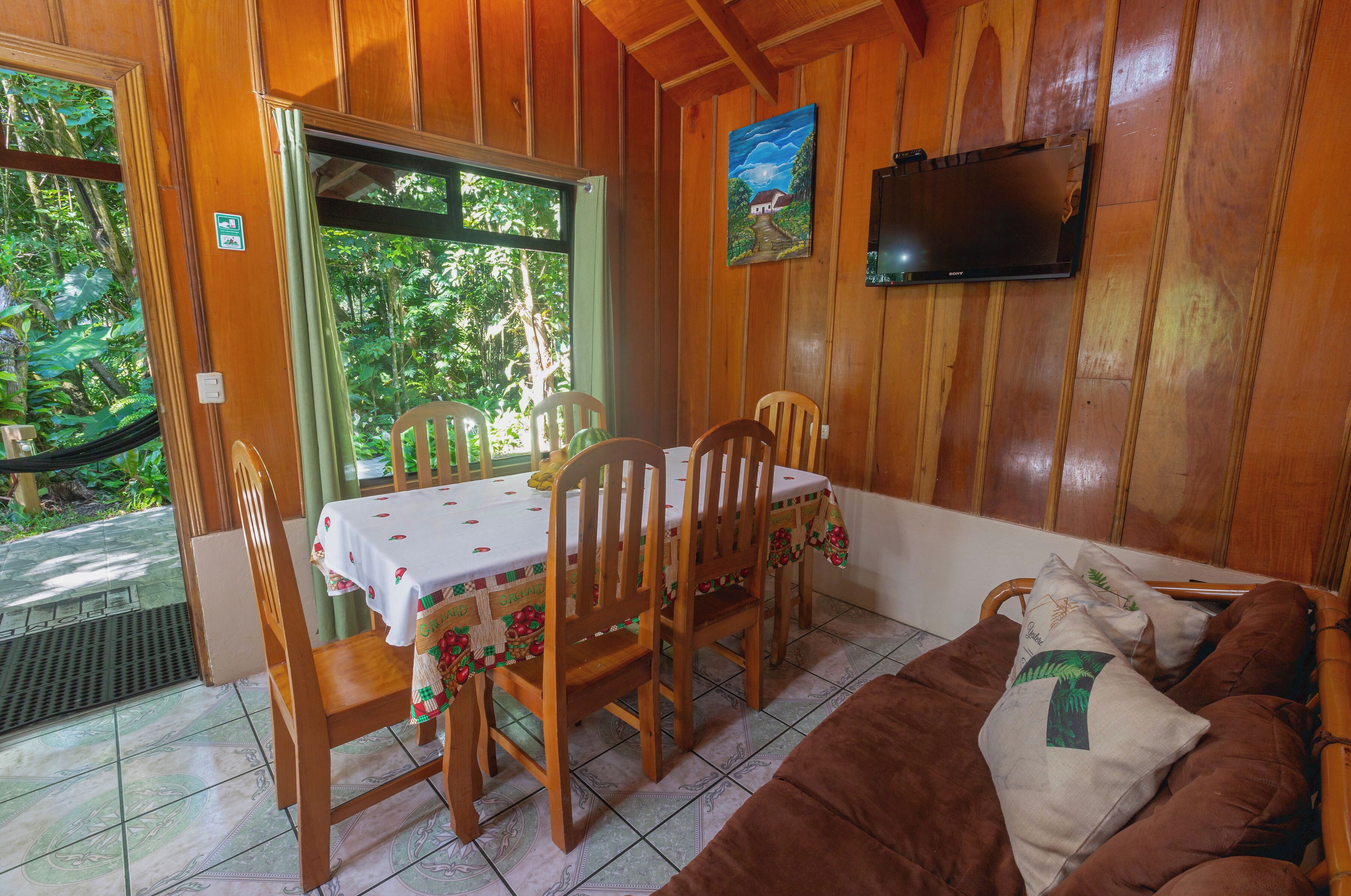 Una habitación con paneles de madera, una mesa de comedor con sillas, un sofá marrón y un televisor montado en la pared.