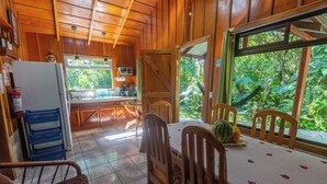 A wooden cabin interior with a dining area, kitchen, and large windows overlooking a lush garden.