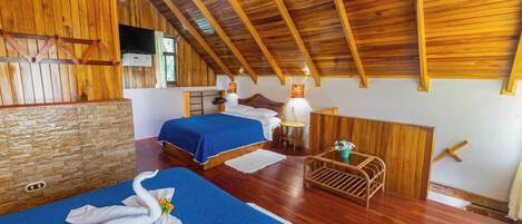Una habitación con suelo de madera, dos camas, un banco de madera y una mesita con lámpara.