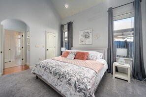 Un dormitorio con una cama grande, una mesita de noche, una ventana con cortinas y una puerta que da a otra habitación.
