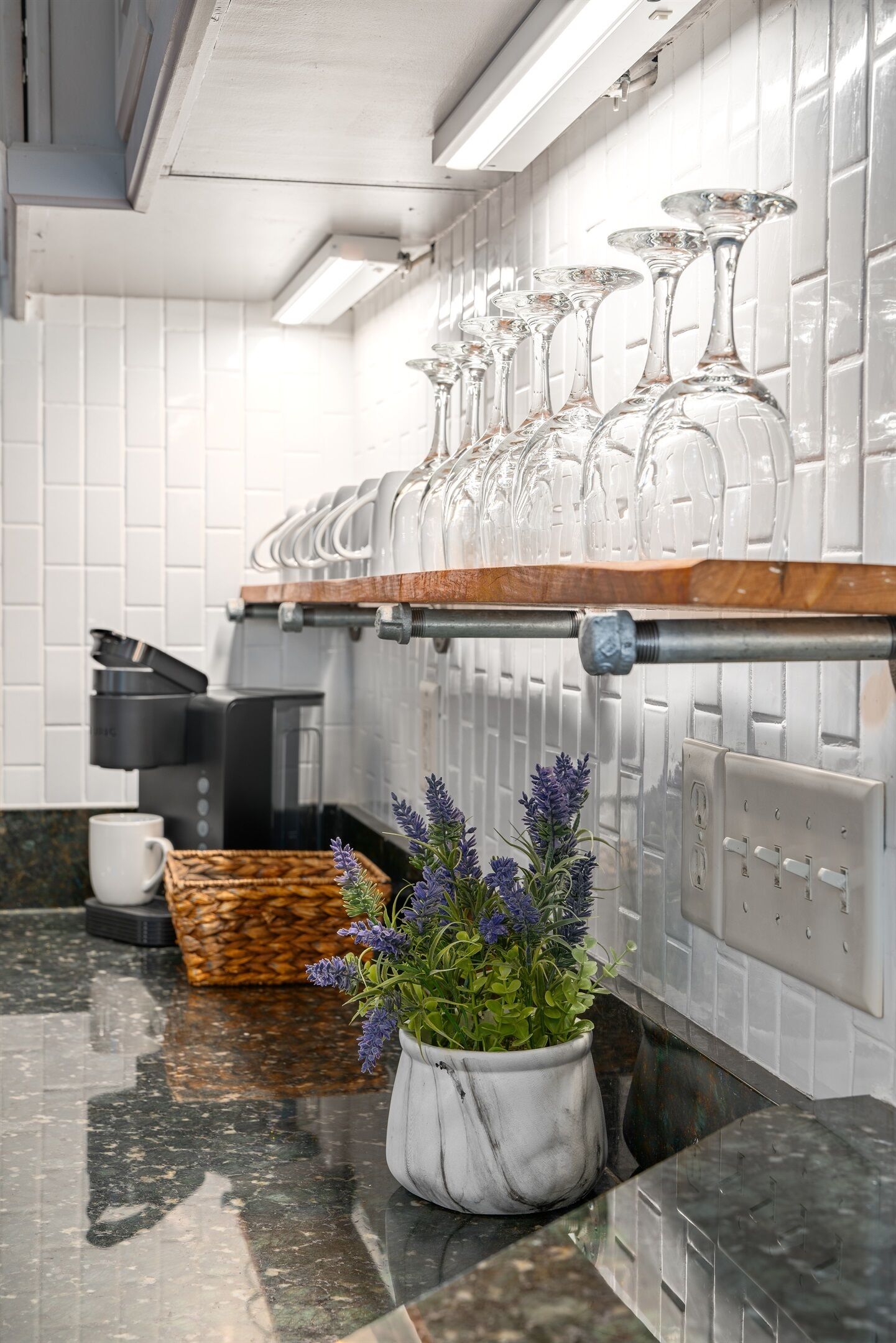 Una cocina con encimera de mármol, una cafetera, una taza blanca, una cesta y una planta en maceta con flores moradas.