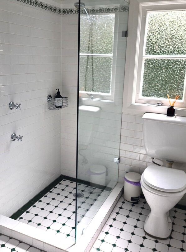 A bathroom with white tiled walls, a glass shower enclosure, a white toilet, and a window with a green patterned view. - Mildenhall - an elegant country 6 bedroom home in beautiful Bundanoon NSW (Bundanoon)