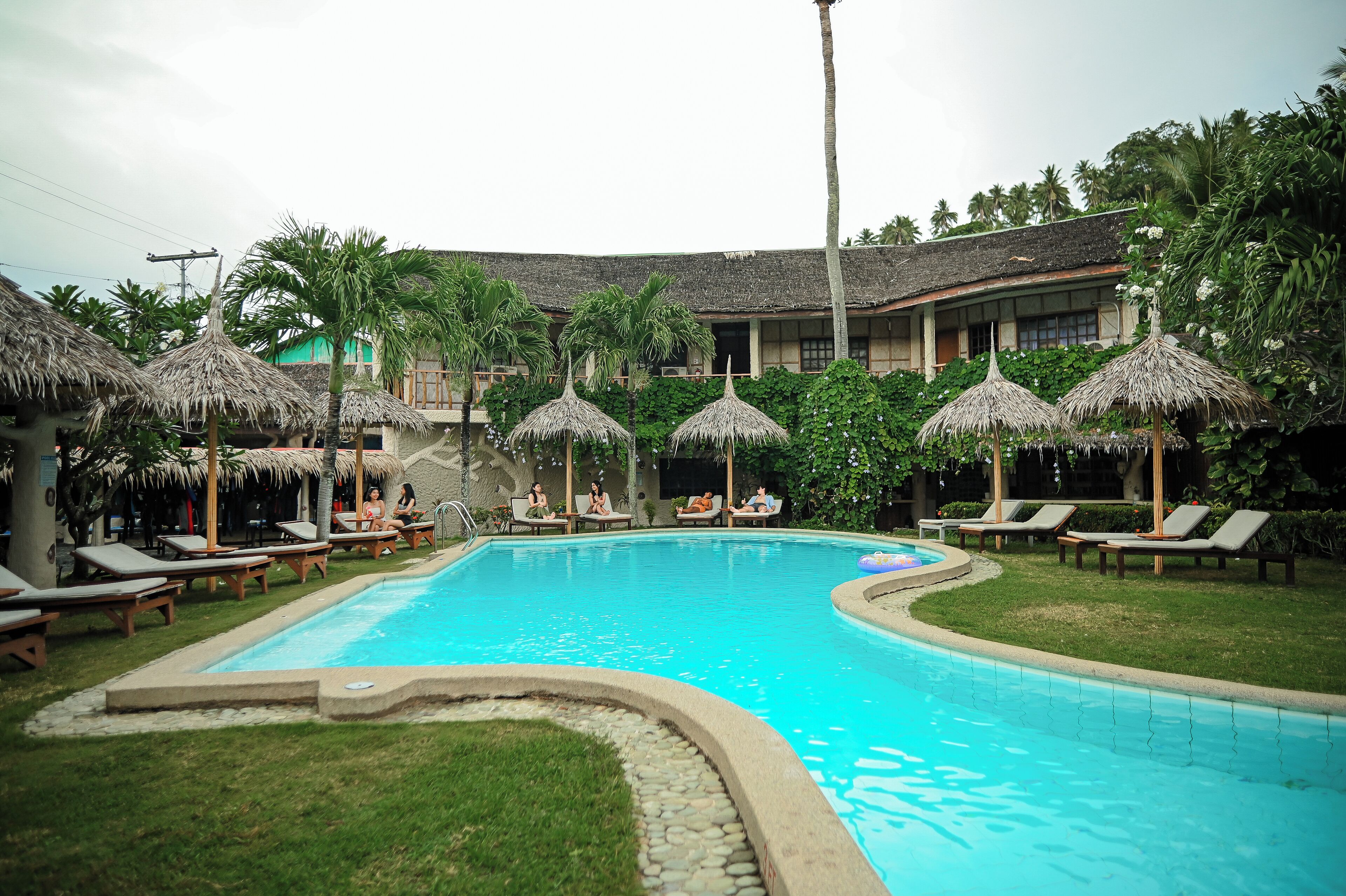 A swimming pool with lounge chairs and thatched umbrellas, surrounded by lush greenery and a building with a tiled roof.
