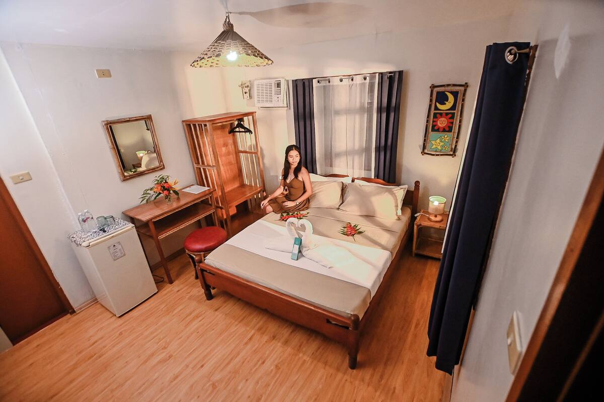 A hotel room with a bed, a desk, a chair, a small refrigerator, a hanging light fixture, and a wall-mounted air conditioner.