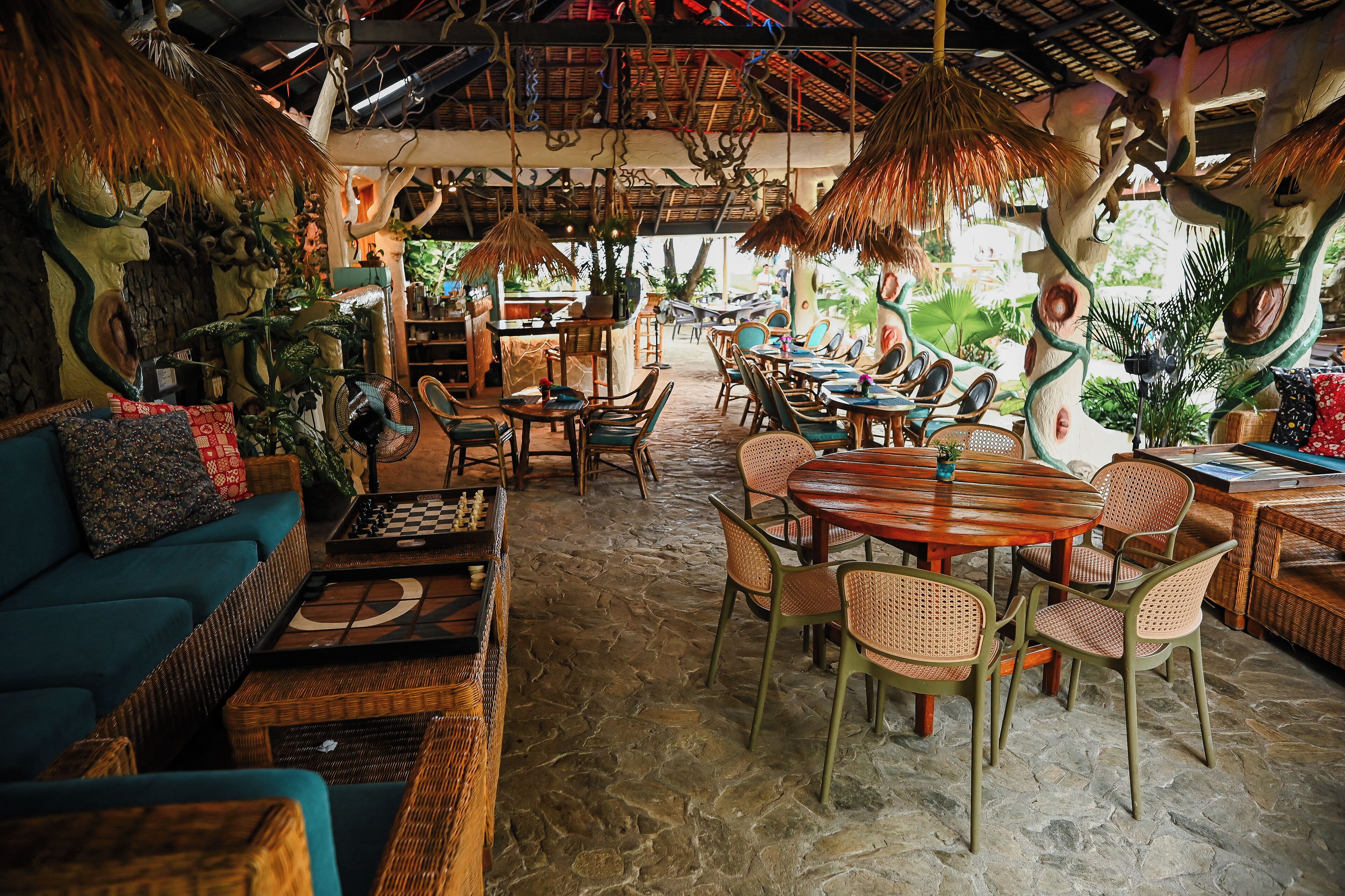 Un restaurant chaleureux, à l’ambiance tropicale, avec des meubles en osier, un jeu d’échecs et un toit de chaume.