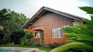 Una casa de madera con balcón, rodeada de abundante vegetación y una piscina.