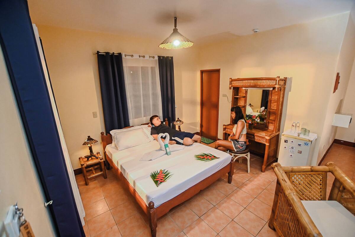 A bedroom with a bed, a desk, a chair, a mirror, and a refrigerator.