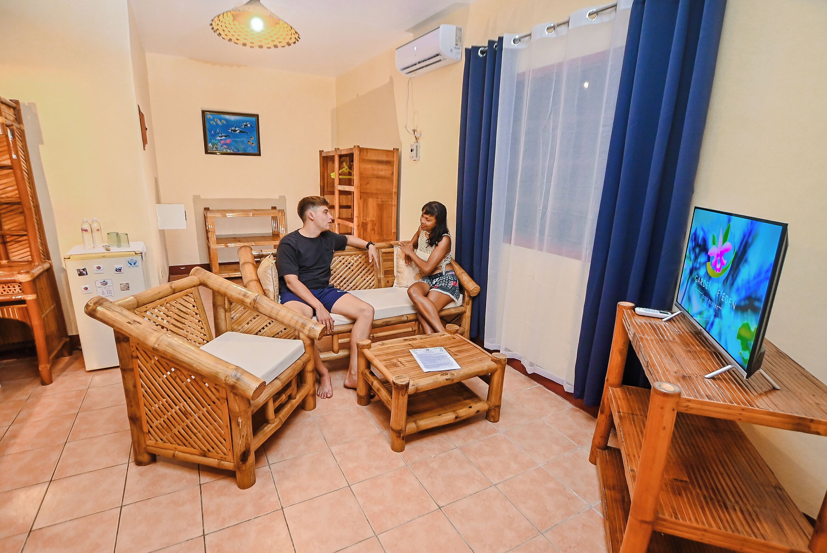 Una habitación con dos sofás de mimbre, una pequeña mesa de madera, un televisor sobre un soporte y una ventana con cortinas.