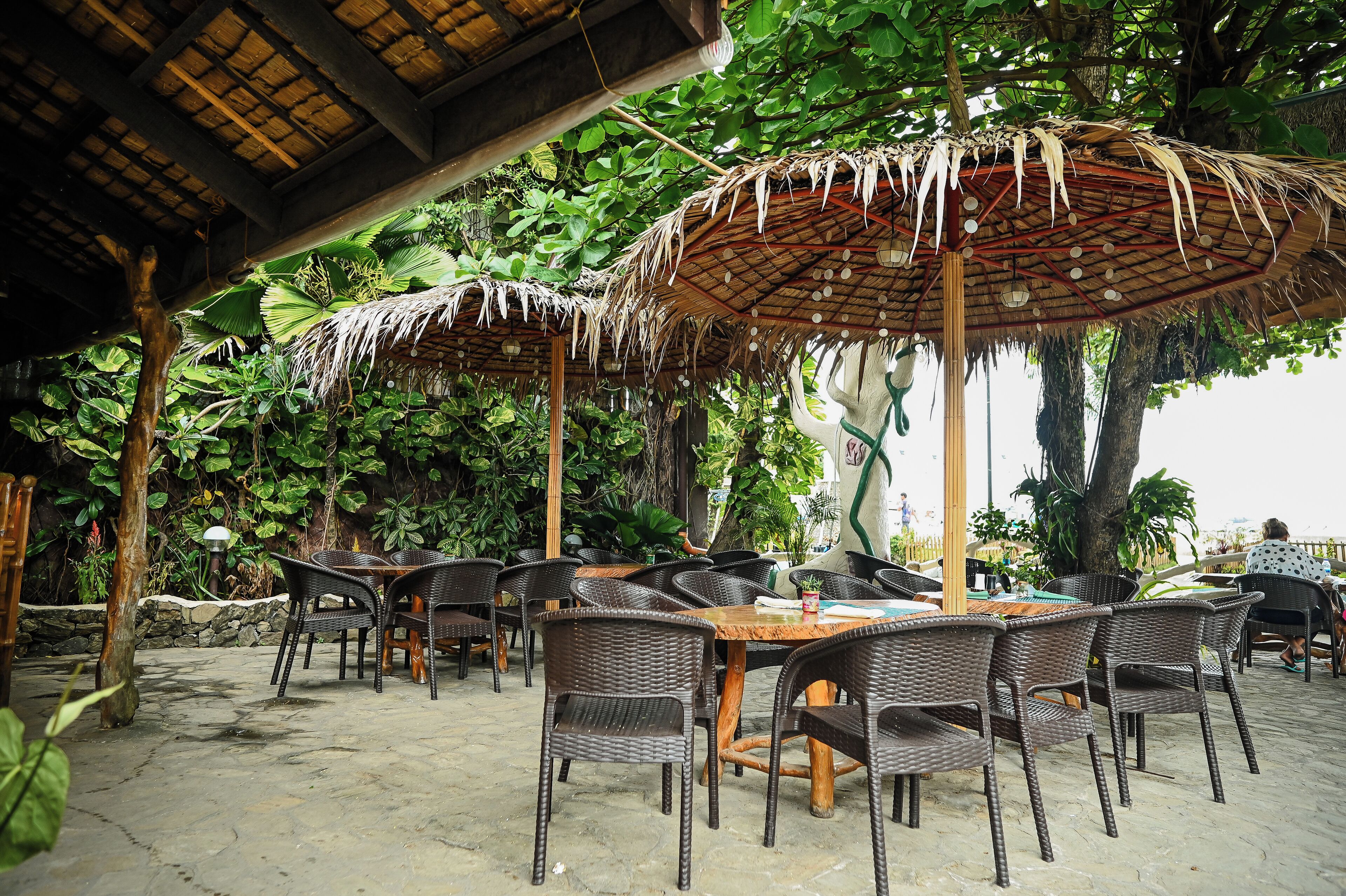 An outdoor seating area with wicker chairs and tables under thatched umbrellas, surrounded by lush greenery.