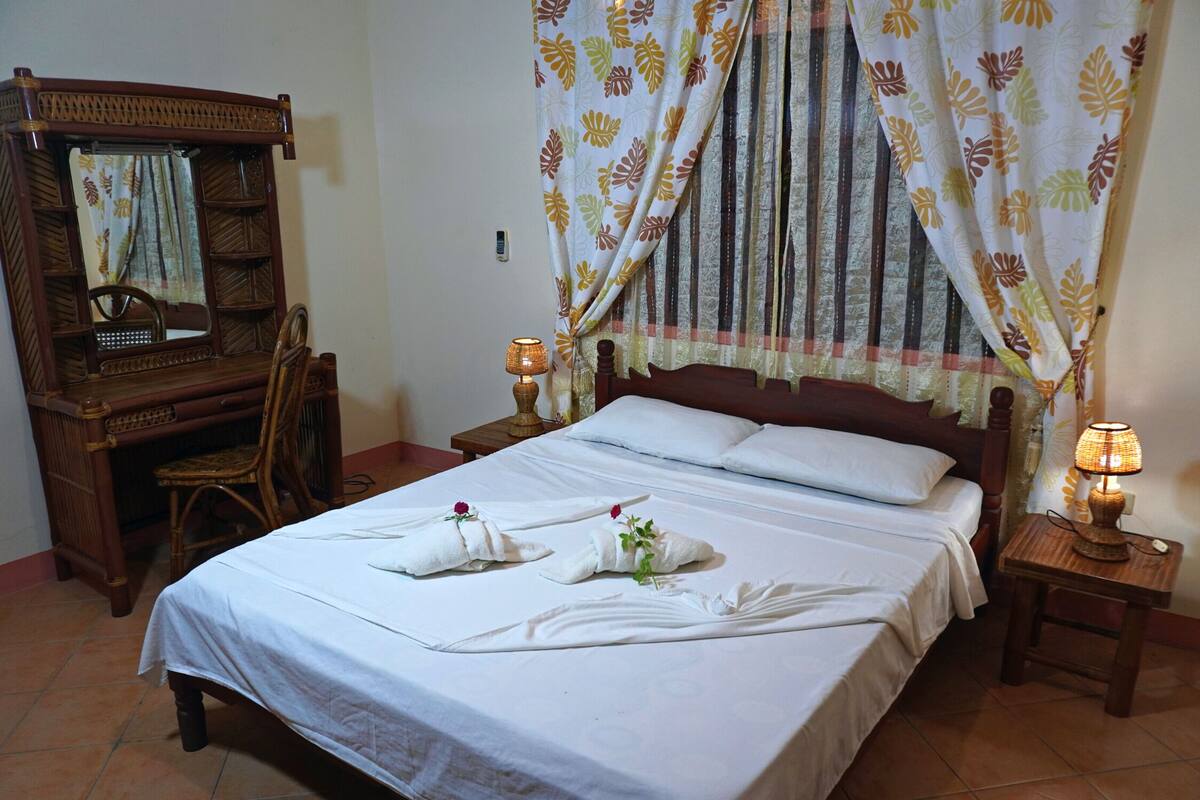 A neatly made bed with two towels and a flower, a wooden desk with a chair, a bedside lamp, and a curtain with a leaf pattern.