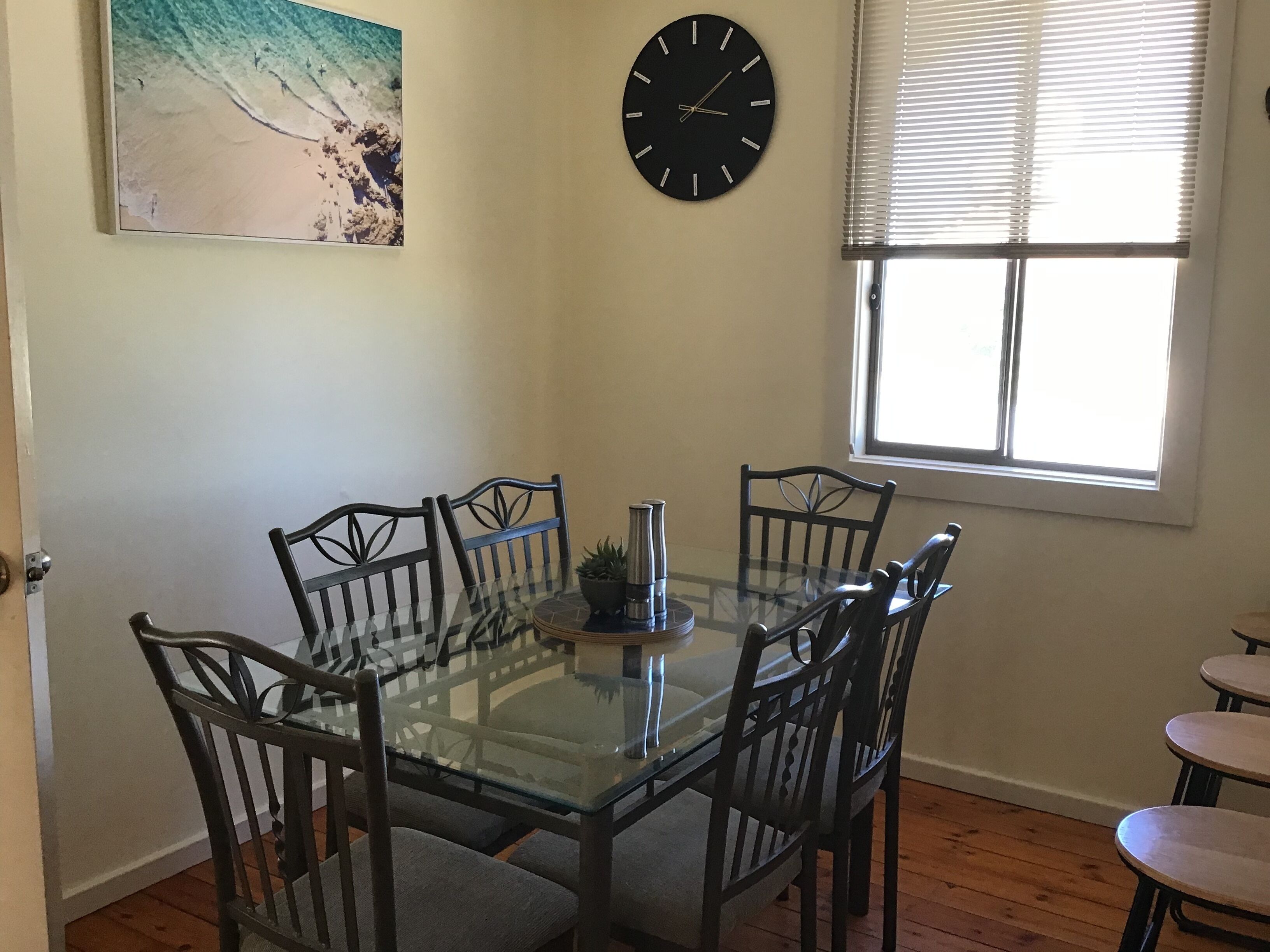 A dining area with a glass-top table, six chairs, a clock, a window with blinds, and a beach-themed painting.