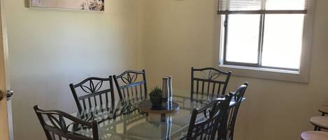 A dining area with a glass-top table, six chairs, a clock, a window with blinds, and a beach-themed painting.