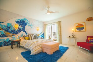 Une chambre avec un décor mural représentant des vagues, un lit avec un tapis bleu, un ventilateur de plafond et une chaise rouge.