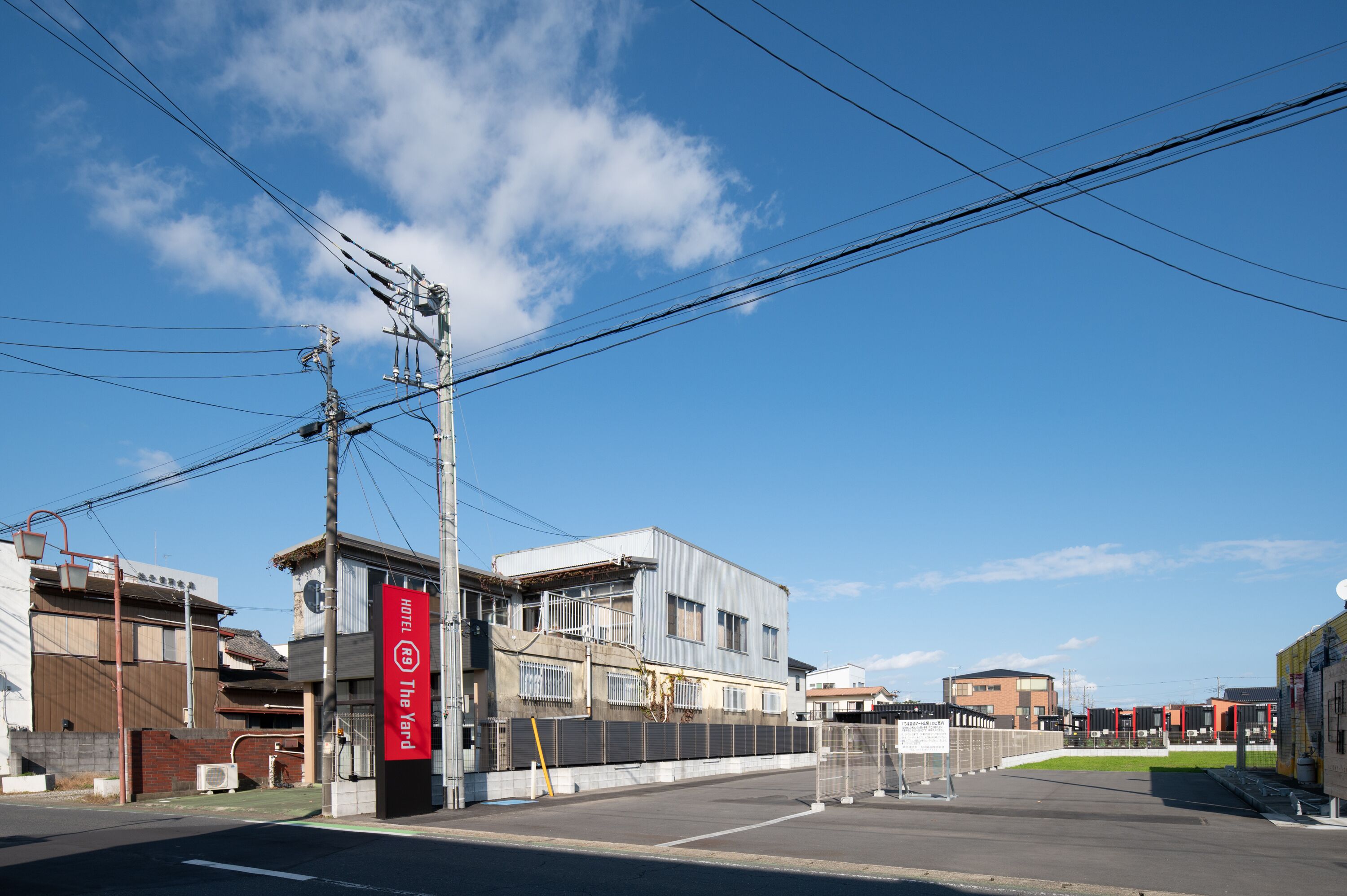 A hotel with a sign reading "HOTEL RE The Yard" in front of a building with a fenced parking area and a clear blue sky.