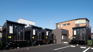 Three black train car containers with numbers 08, 09, and 29, parked in a lot with a modern building in the background. - HOTEL R9 The Yard Asahishiro (Asahi)
