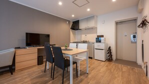 A modern kitchen with white cabinets, a built-in oven, and a microwave. There is a dining area with a table and chairs, and a TV on a stand. - HOTEL R9 The Yard Asahishiro (Asahi)