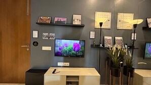 Zona de recepción de un hotel con un televisor mostrando una imagen floral, un mostrador de recepción y varias fotografías enmarcadas en la pared.