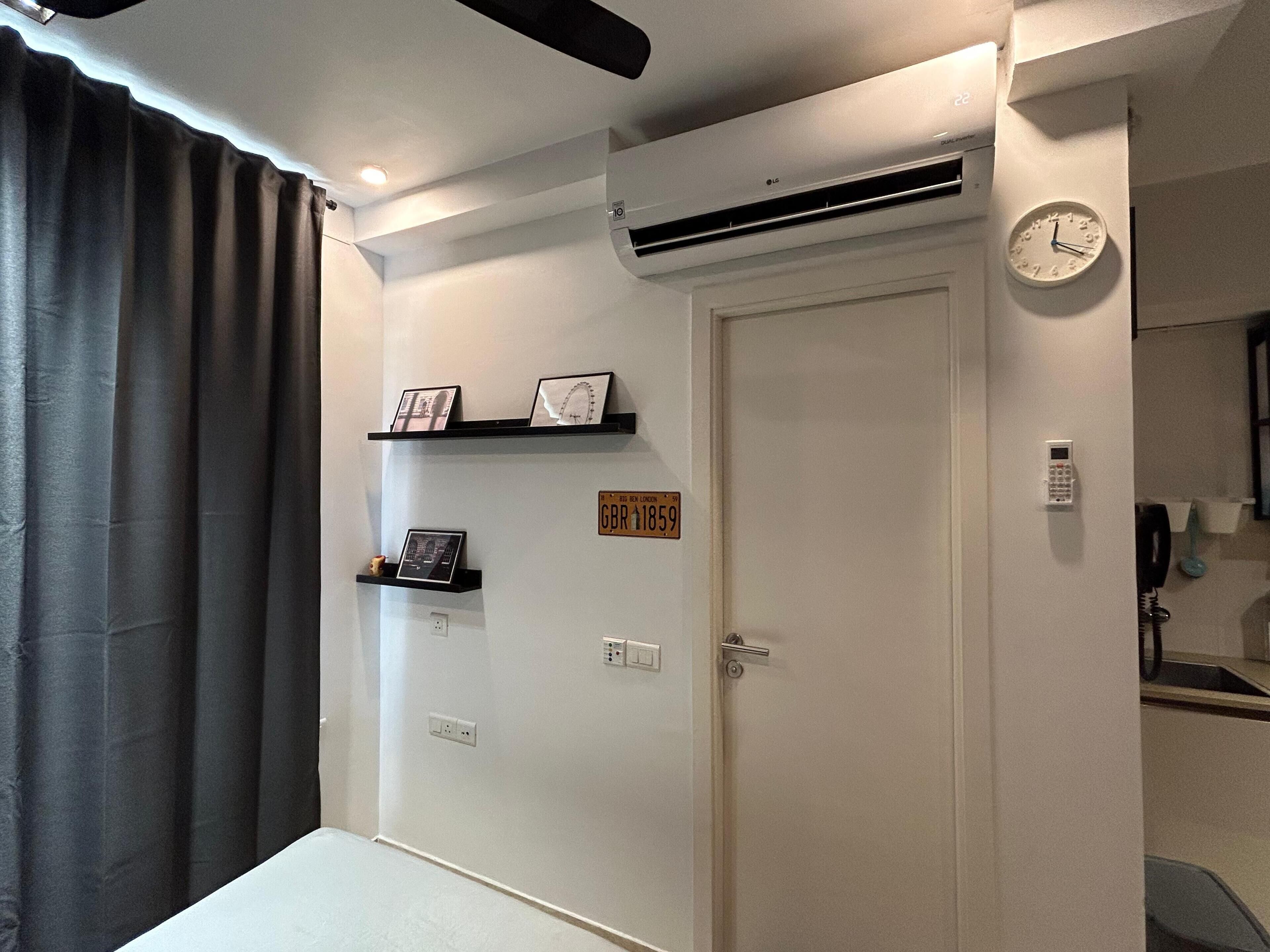 A hotel room with a door, air conditioning unit, a clock, and a shelf with framed pictures.