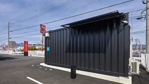 A large, black, metal structure with a flat roof, likely a bus stop or shelter, situated on a paved area with a white line marking. - HOTEL R9 The Yard Tarui (Tarui)