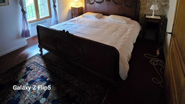 Ein Schlafzimmer mit Holzbalkendecke, einer Steinwand, einem großen Bett mit Holzfußteil, zwei Nachttischen mit Lampen und einem gemusterten Teppich.