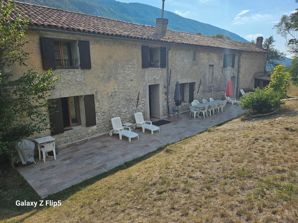 Un bâtiment rustique en pierre, doté d’un toit en tuiles, d’un espace extérieur aménagé avec des chaises et des tables blanches, et offrant une vue sur les montagnes en arrière-plan.