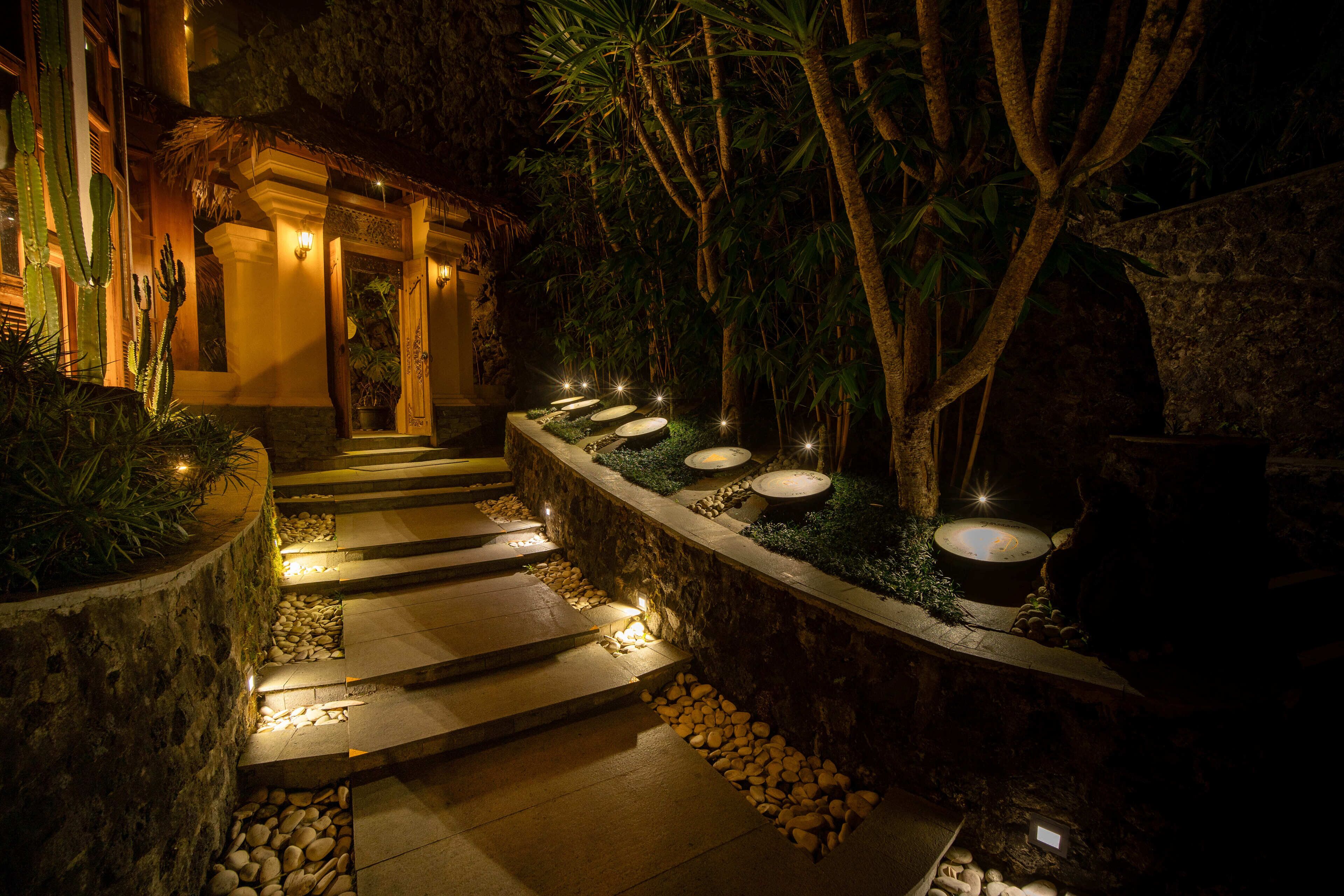 Un sendero de piedra conduce a la entrada de un edificio, rodeado de plantas tropicales e iluminado por luces en el suelo.