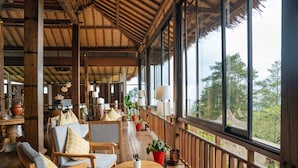 A wooden deck with seating and potted plants, large windows with a view of trees, and a thatched roof. - TIRA VILAGNA SUITES & SPA (Kintamani)
