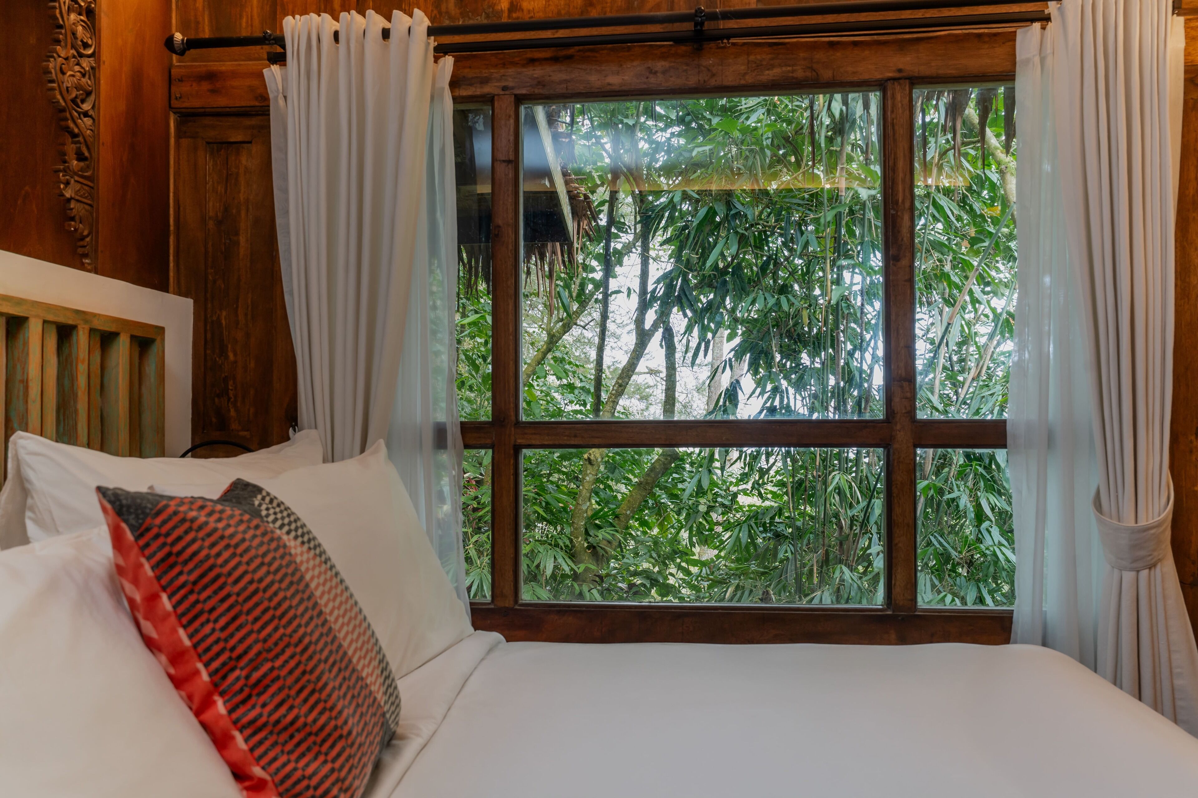 A bedroom with a large window, wooden walls, and a bed with white and patterned pillows.