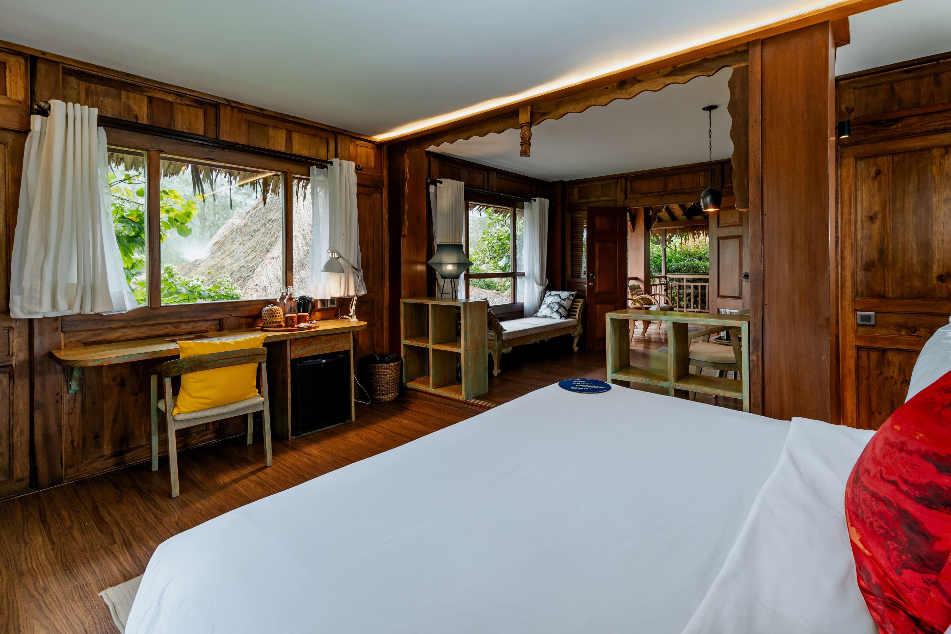 A wooden room with a bed, a desk, and a chair. There is a window with curtains and a view of greenery outside.