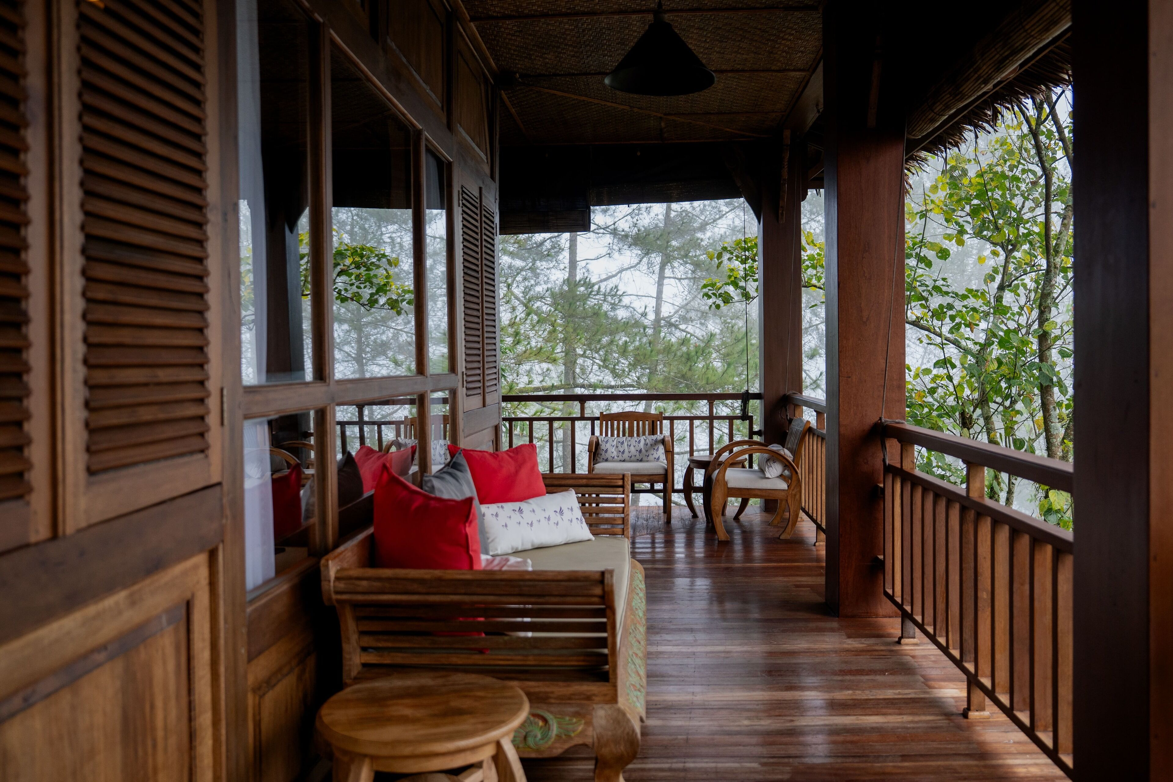 A wooden porch with a sofa and chairs, a railing, and a view of trees through the windows.