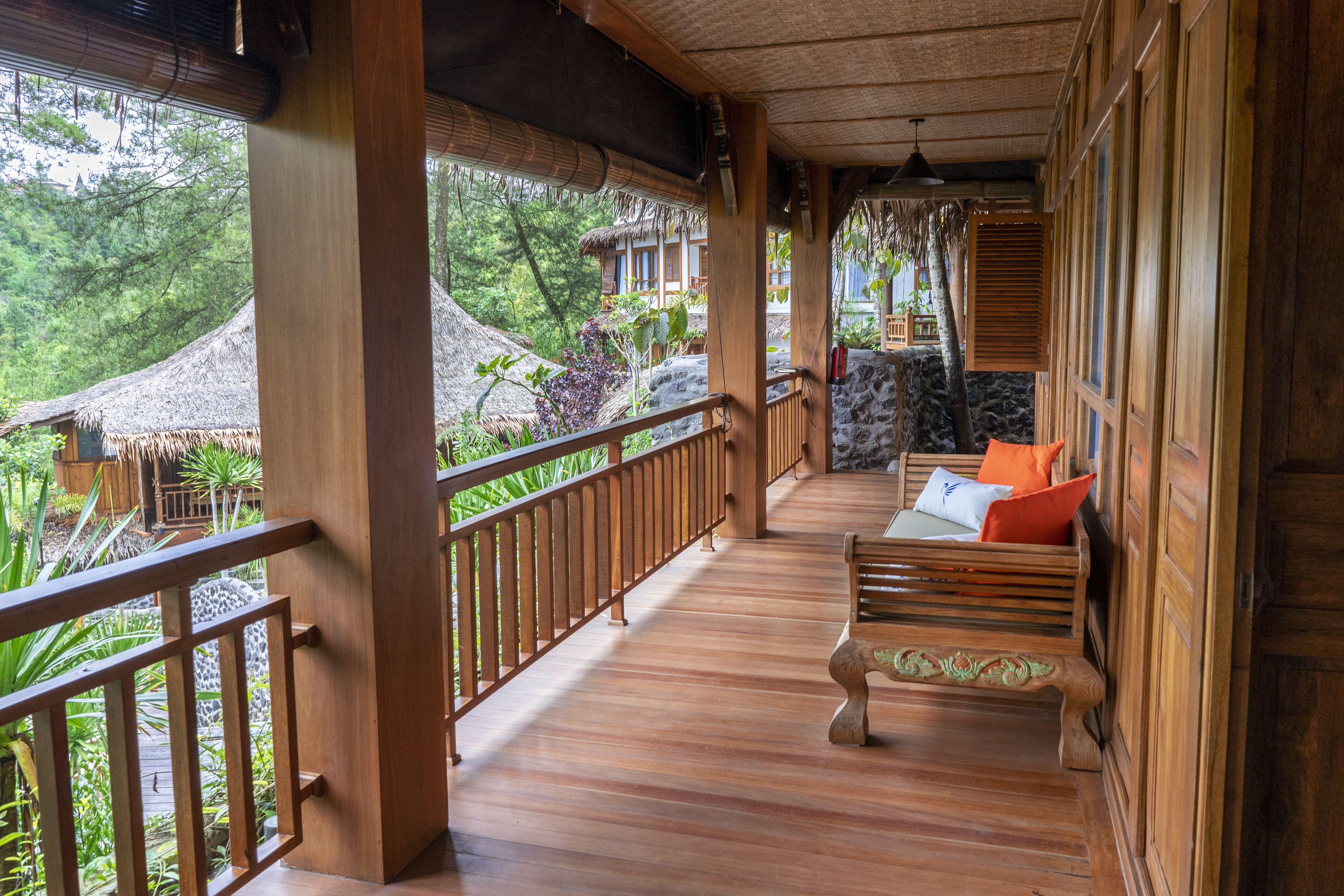 Un balcon en bois avec un banc, une balustrade et une vue sur une construction à toit de chaume et sur la verdure.