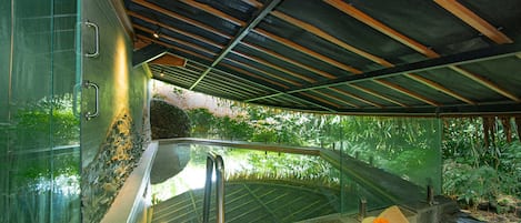 A modern bathroom with a glass enclosure, a wooden bench with towels, and a view of greenery outside.