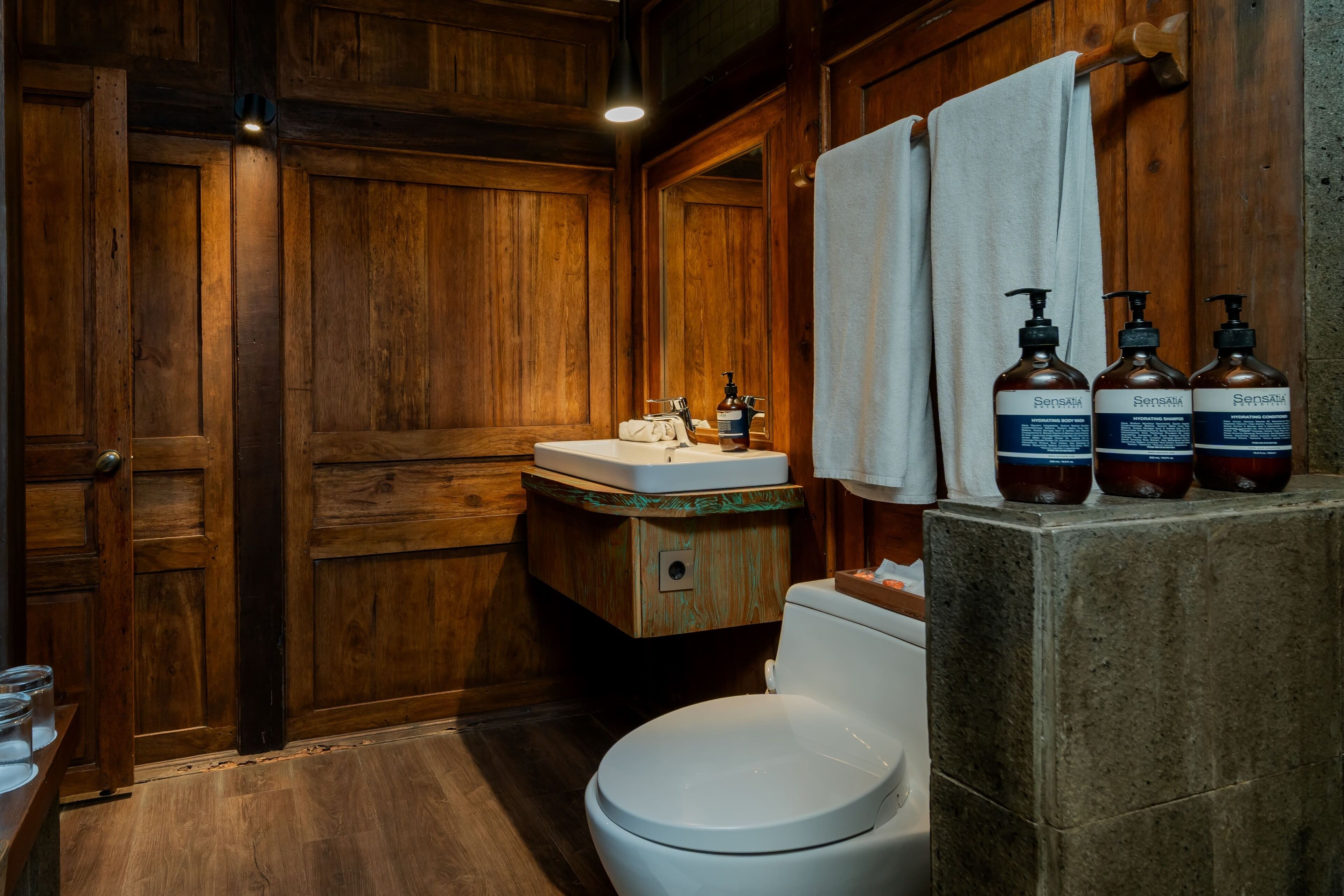 Une salle de bain en bois avec des toilettes blanches, un meuble-lavabo en bois et trois bouteilles de produits Sensatia posées sur le comptoir.