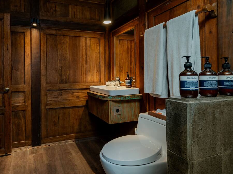 Une salle de bain en bois avec des toilettes blanches, un meuble-lavabo en bois et trois bouteilles de produits Sensatia posées sur le comptoir.