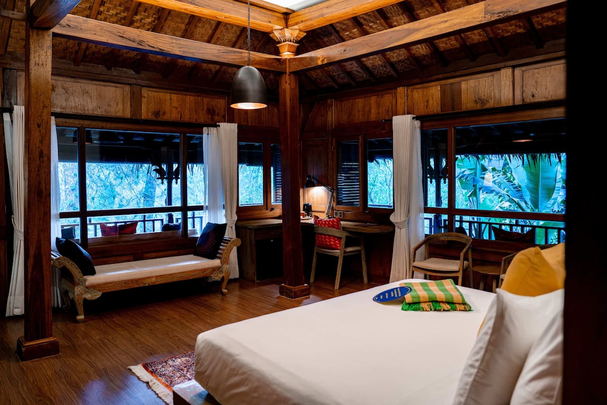A wooden room with a bed, a desk, and a chair. There are windows with curtains and a view of trees.
