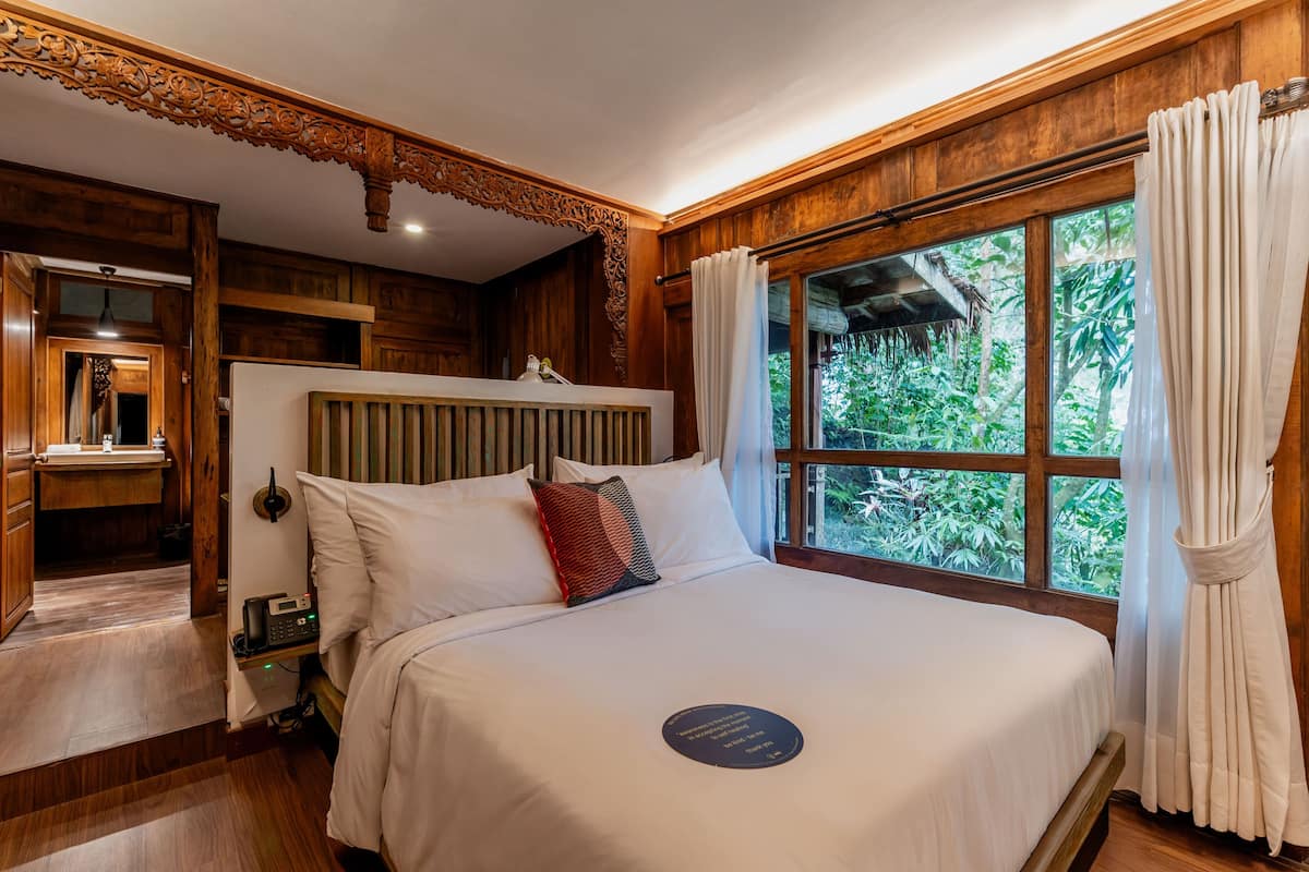 A bedroom with a large bed, wooden headboard, a window with curtains, and a view of greenery outside.