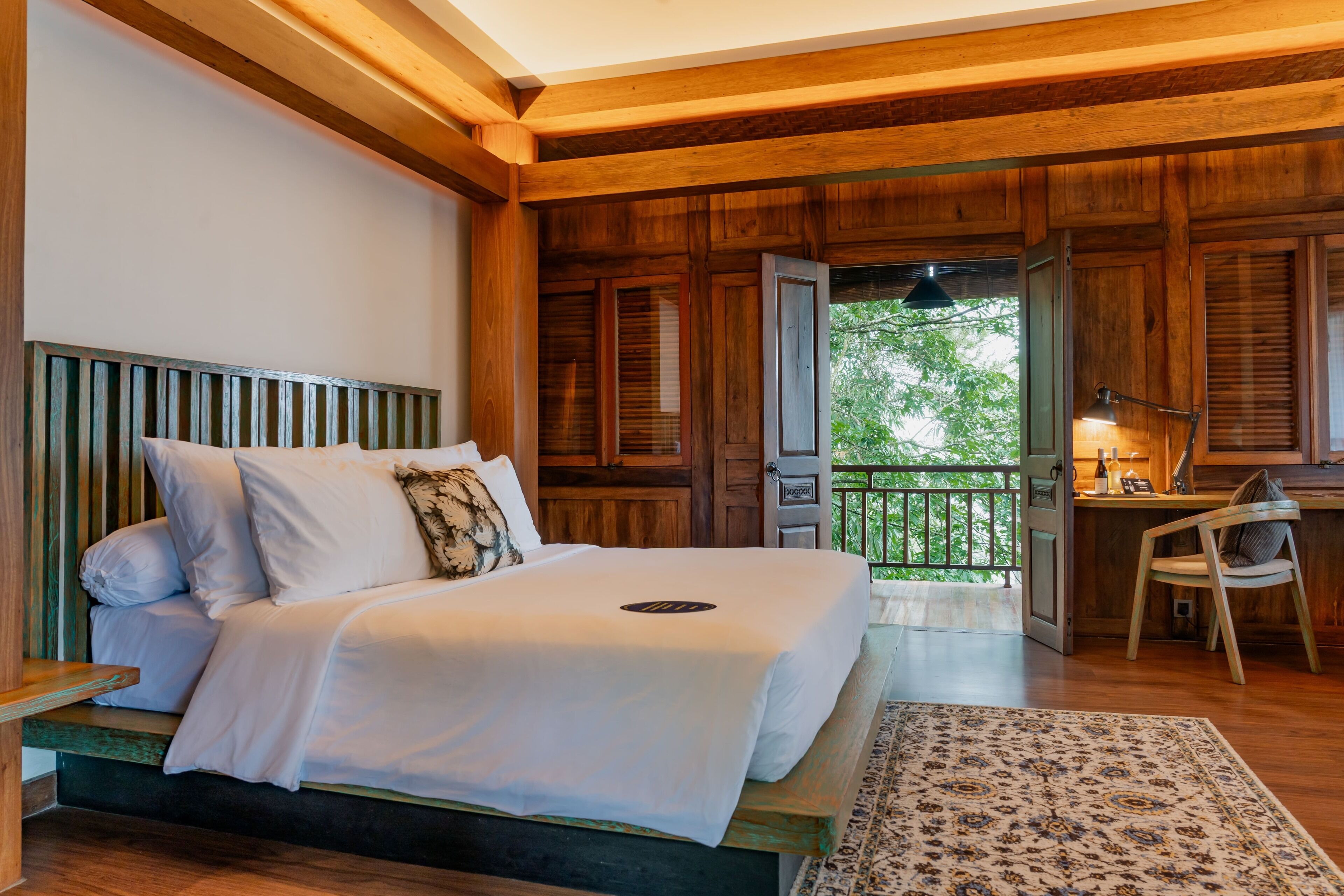 A bedroom with a wooden headboard, a bed with white bedding, a desk with a chair, and a view of greenery outside.