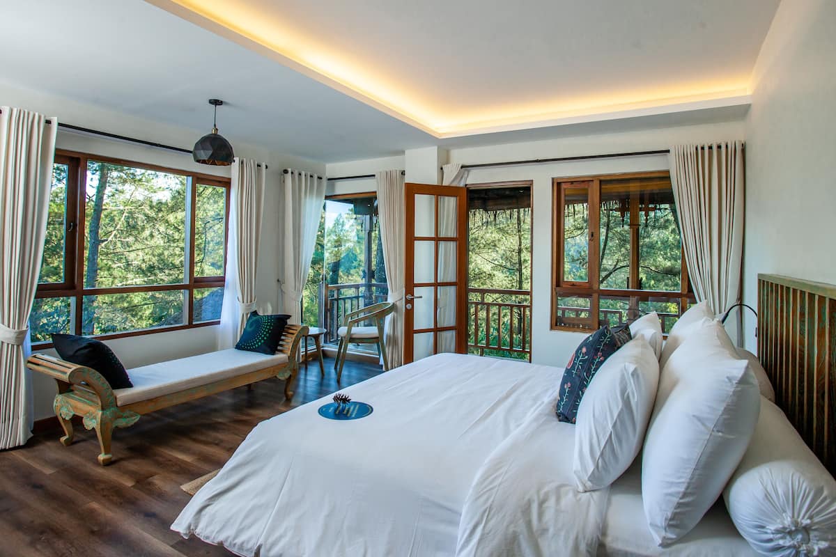 A bedroom with a large bed, a bench with cushions, and a view of trees through the windows.