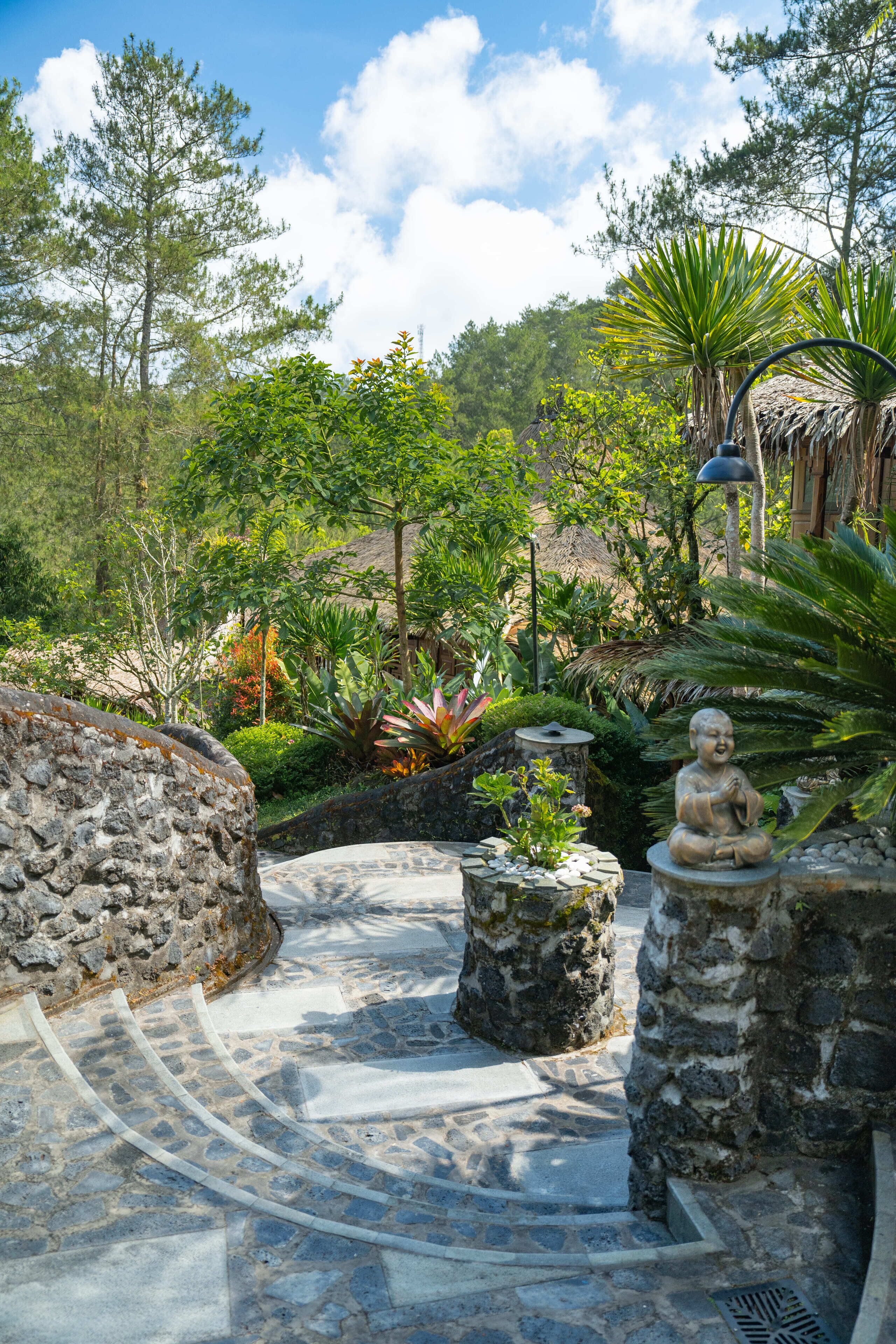 Un sendero de piedra que conduce a una estructura con techo de paja rodeada de plantas tropicales y una estatua de Buda.