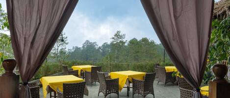 An outdoor dining area with tables and wicker chairs, covered by curtains, and a view of trees and sky.